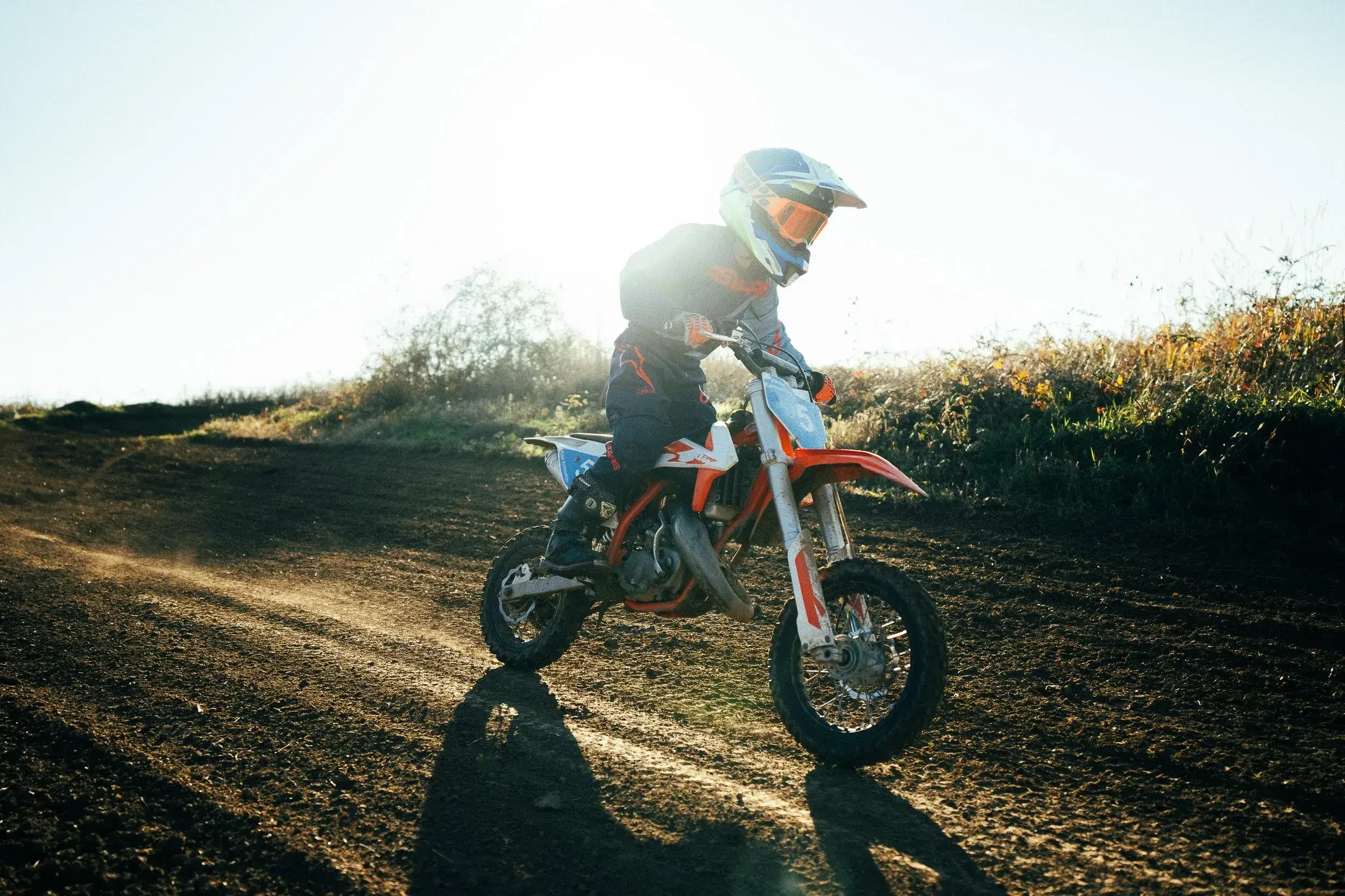 Child riding a dirt bike on a dirt trail during sunset, wearing a helmet and protective gear.

