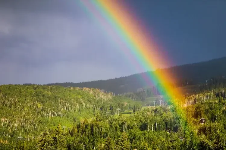 Rainbow arching over a green forested mountain landscape with ski lift structures and a cloudy sky in the background.
