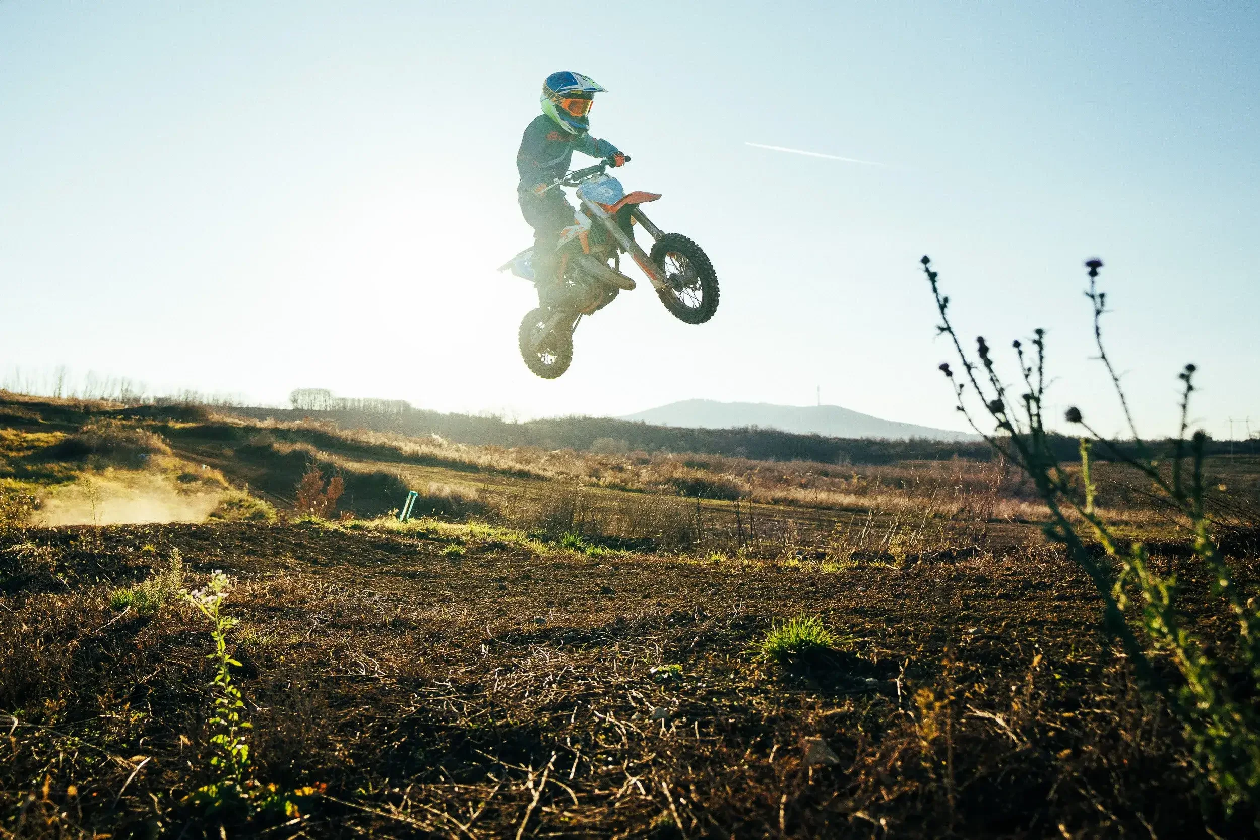 Motocross rider mid-air on orange dirt bike against a clear blue sky, dirt terrain below, in a rural outdoor setting.
