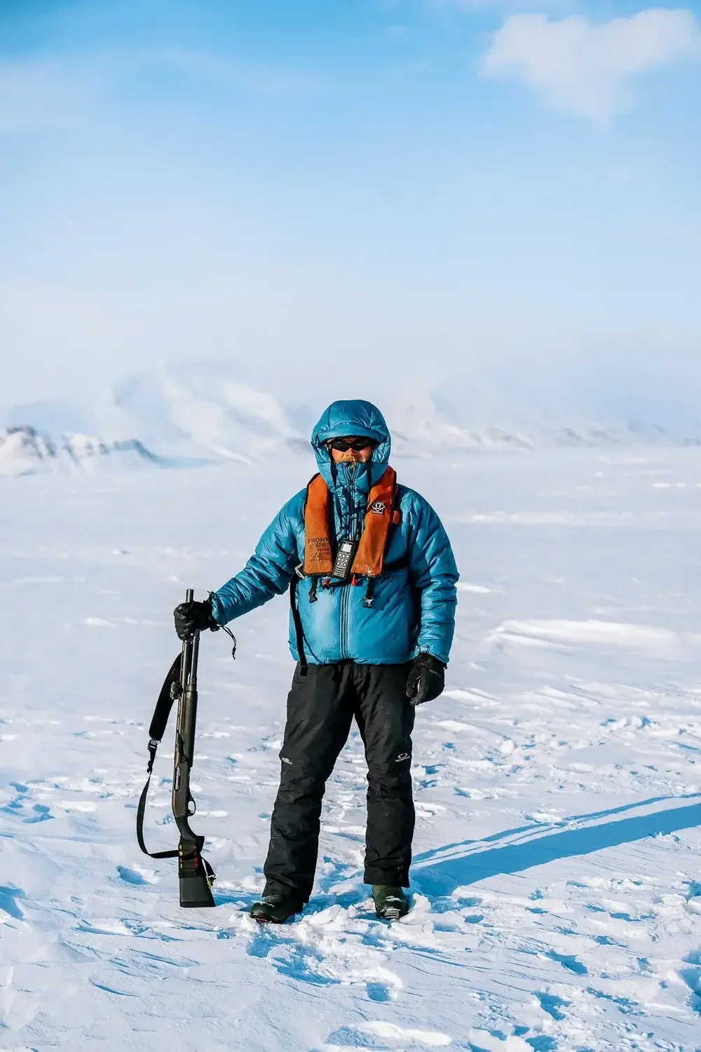 Person dressed in blue winter jacket and black pants standing on snow with a telephoto lens and camera equipment, winter landscape in background.
