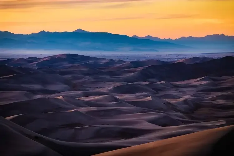Colorful landscape of rolling sand dunes at sunset with distant mountains and a pastel sky.
