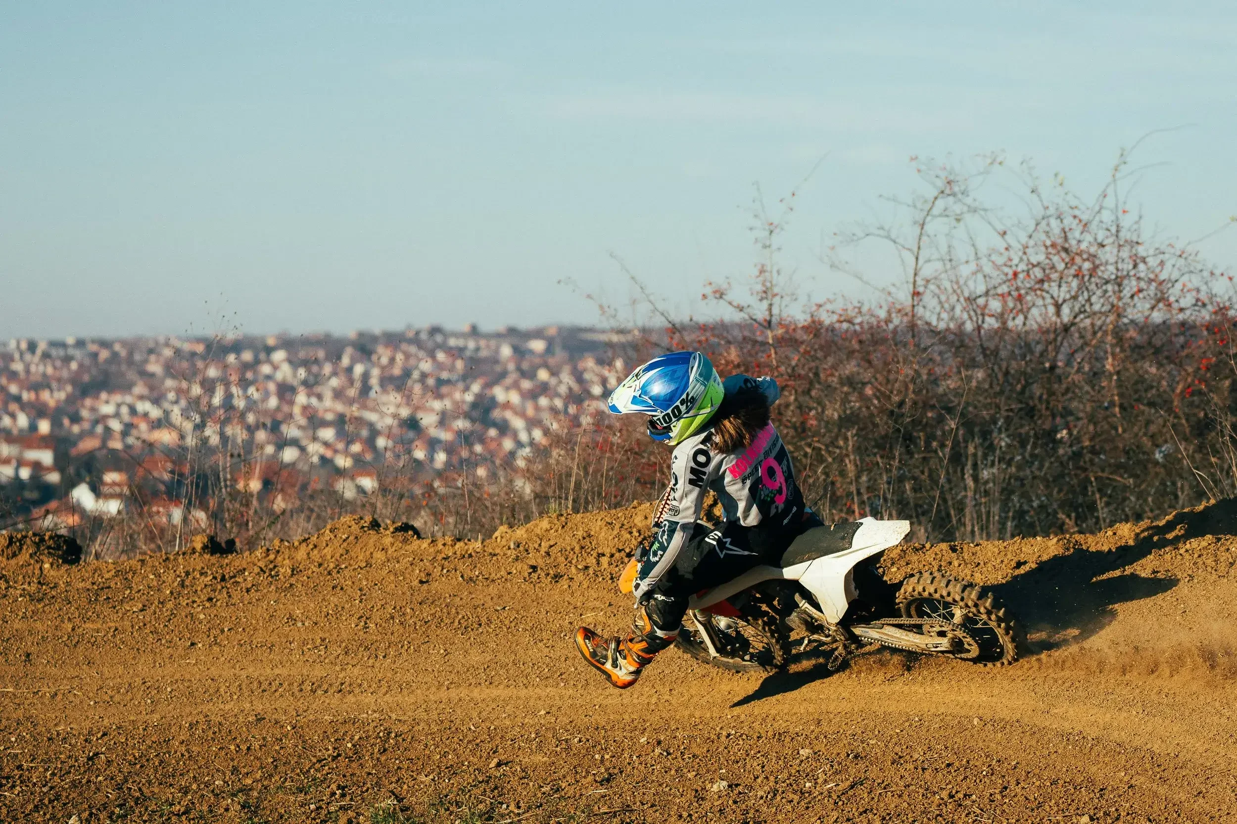 A person riding a dirt bike on a dirt trail, wearing a helmet and protective gear, with a cityscape in the background.
