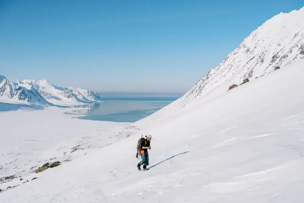 A person hiking up a snowy mountain slope with snow-covered peaks and a body of water in the background on a clear day.
