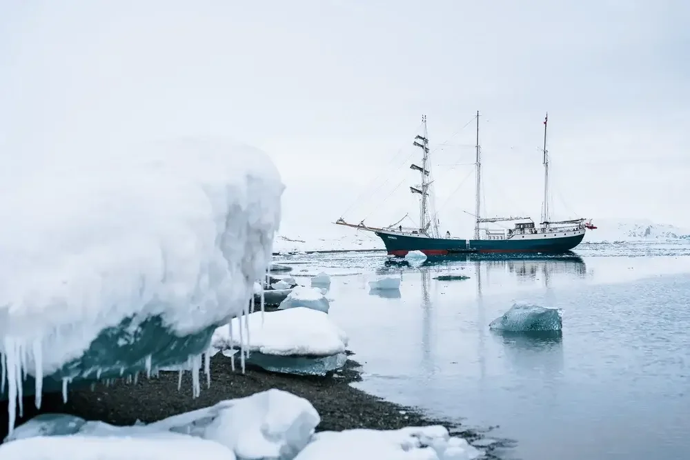 A ship in icy waters near a snow-covered shoreline with ice chunks and snow in a cold, Arctic environment.