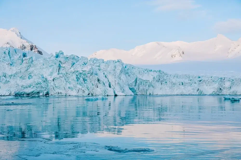 A glacier with ice falling into icy water in a cold, snowy landscape with snow-covered mountains in the background.
