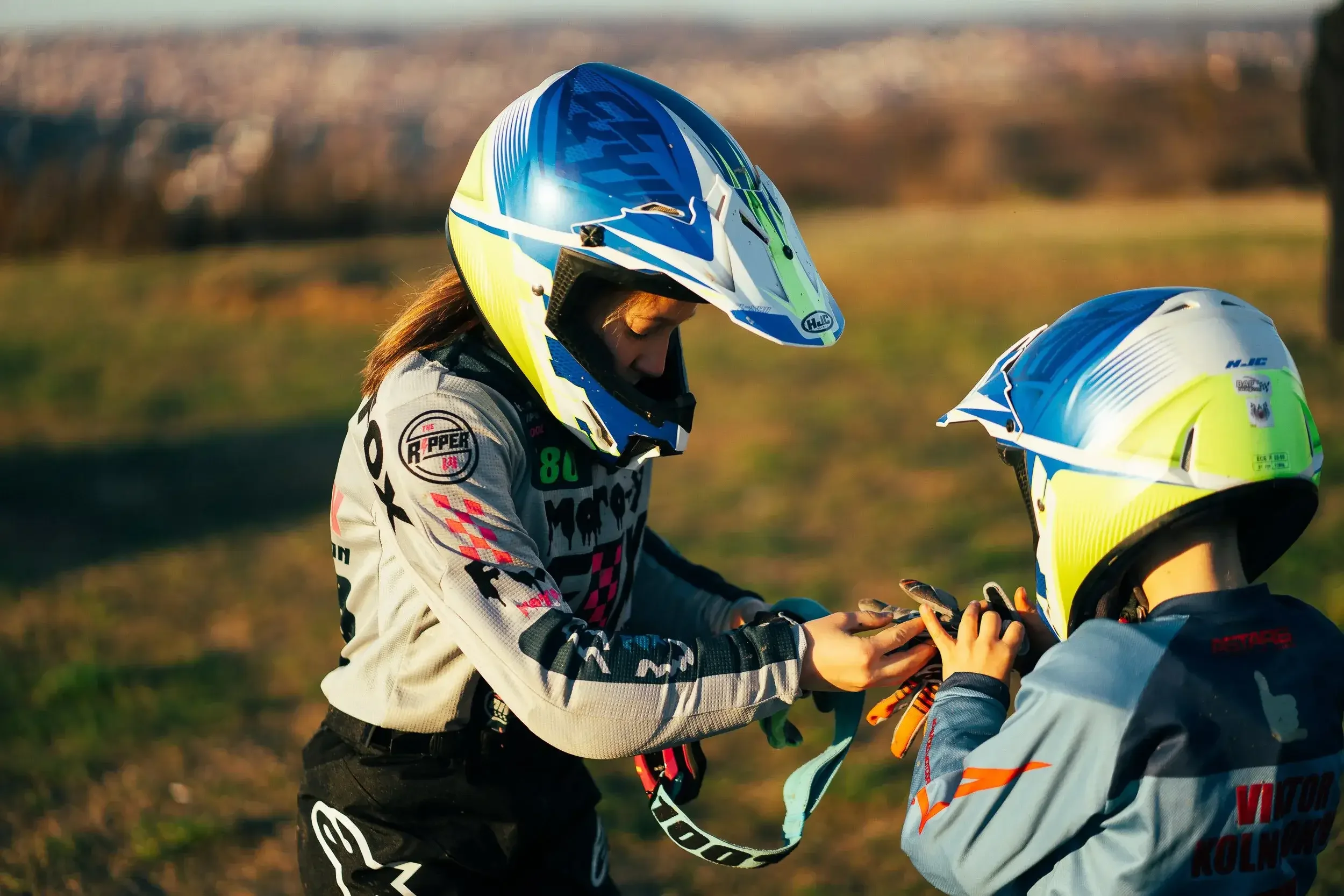 Two young motocross riders wearing helmets and racing gear exchanging goggles outdoors in a grassy area with a blurred background.
