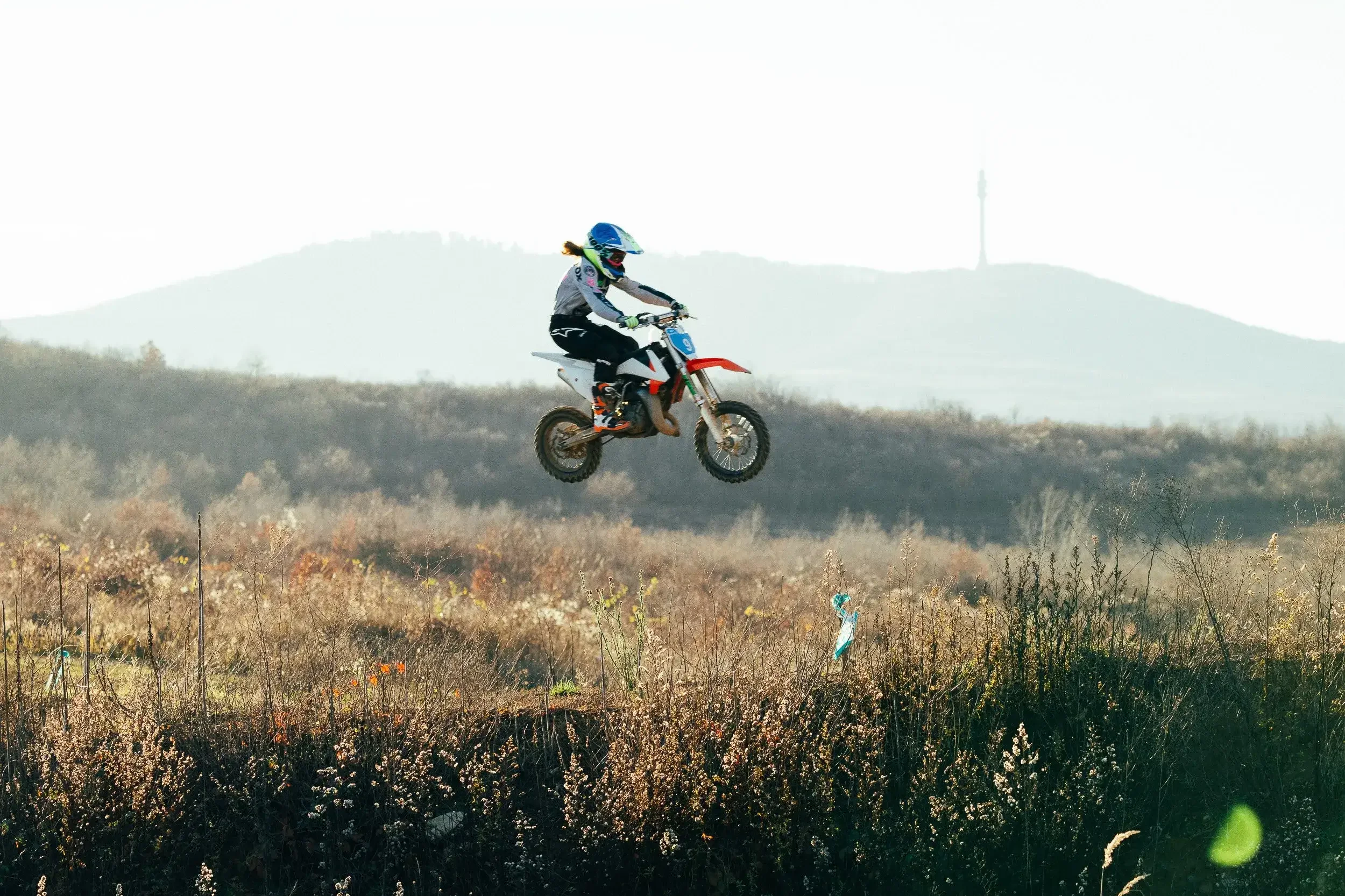 A person in motocross gear jumping a dirt bike in a field with hills and a tower in the background.
