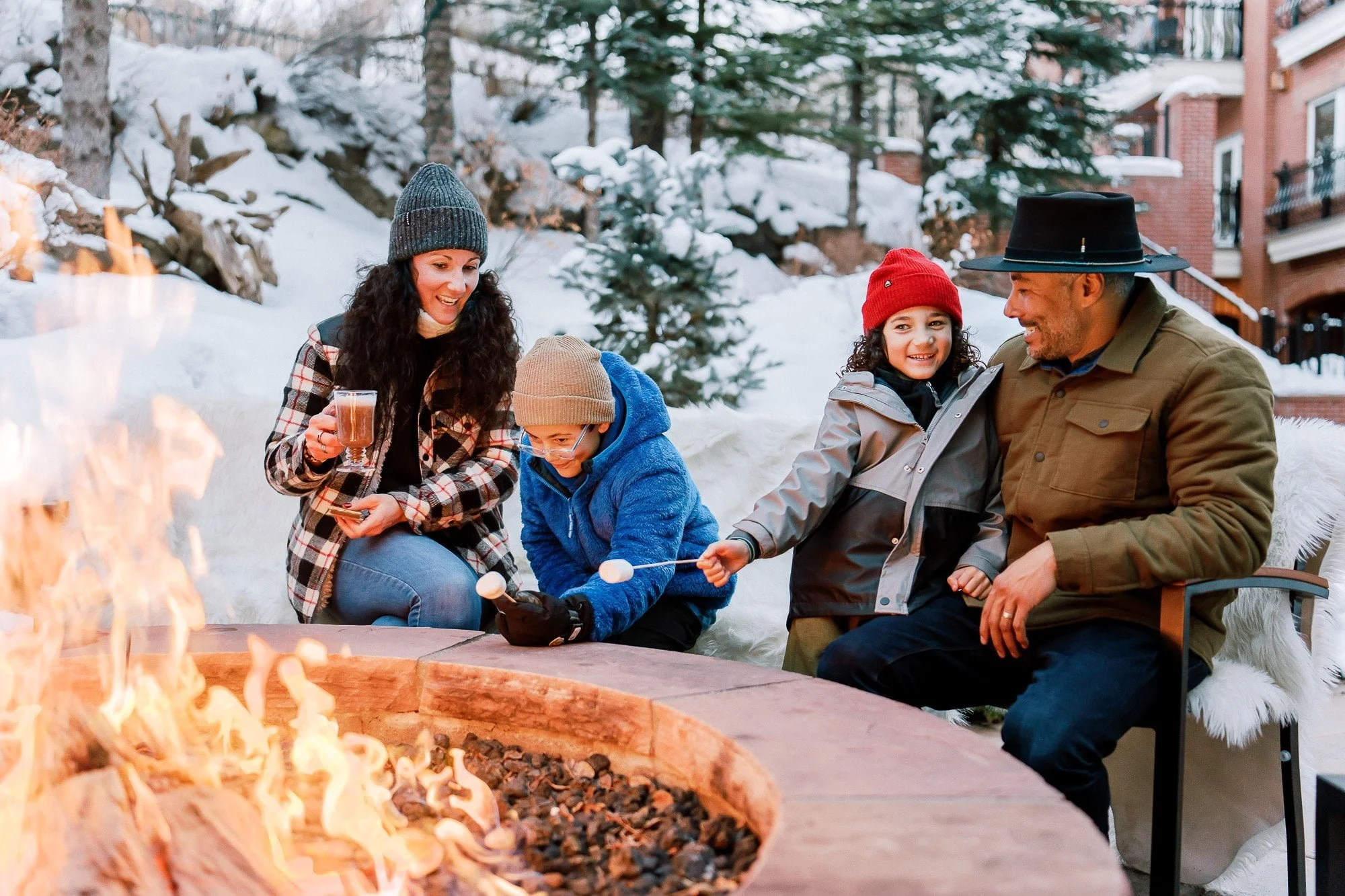 A family of four sitting outdoors around a fire pit in a snowy yard, roasting marshmallows and enjoying drinks together.