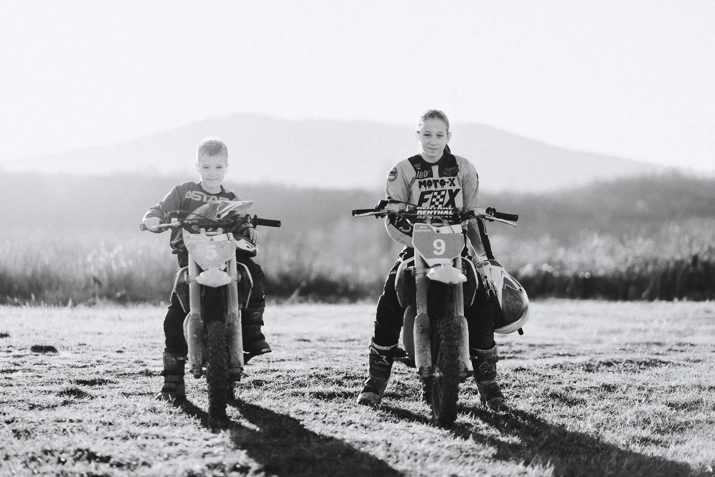 Two children riding dirt bikes on a grassy field with mountains in the background, smiling and looking at the camera, in black and white.

