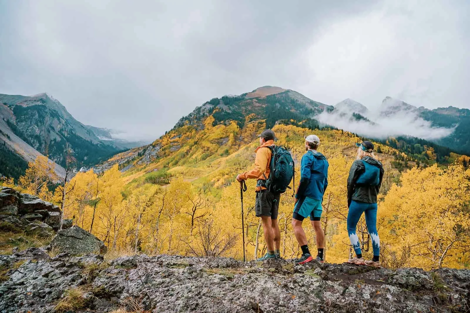 Three hikers with backpacks and outdoor gear standing on rocky terrain, overlooking a scenic mountain landscape with autumn-colored trees and misty clouds.
