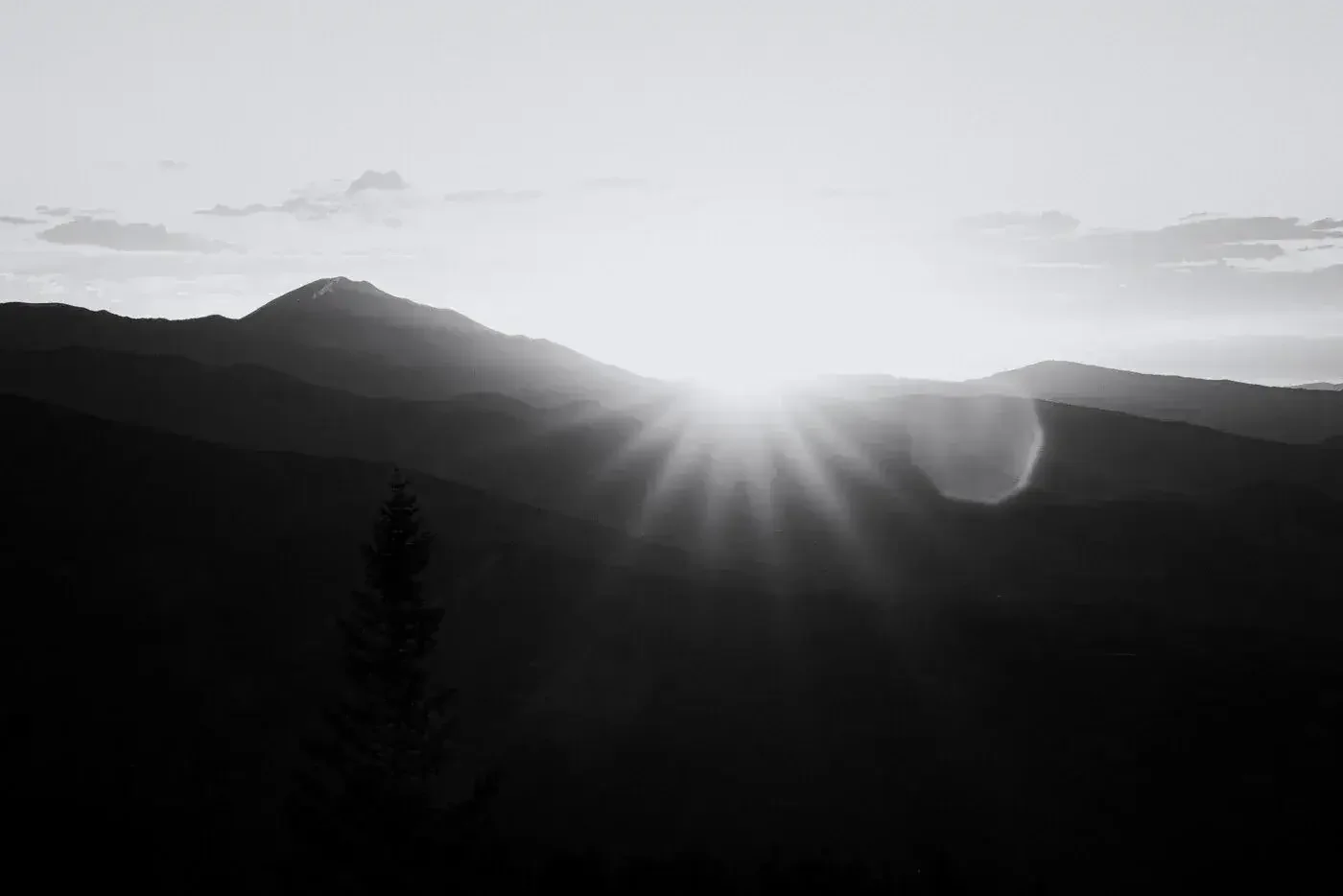 Black and white photograph of a mountain landscape with sunlight shining over the horizon, creating sun rays and lens flare.