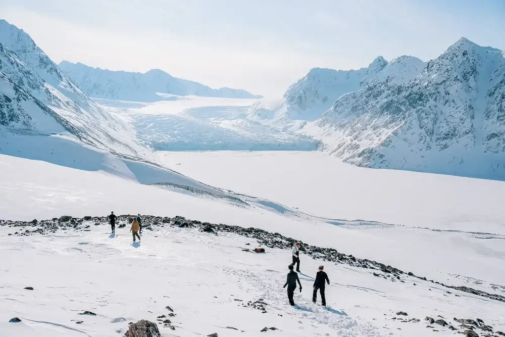Group of people hiking on snowy terrain in a glacier landscape with snow-covered mountains and ice in the background.
