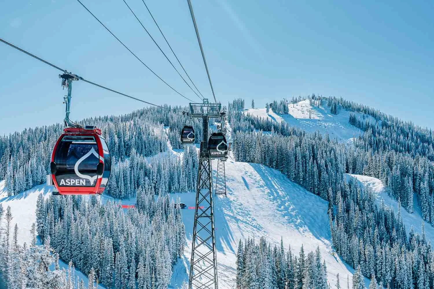 Ski gondola lift carrying passengers at Aspen ski resort, ascending snowy mountain with pine trees.