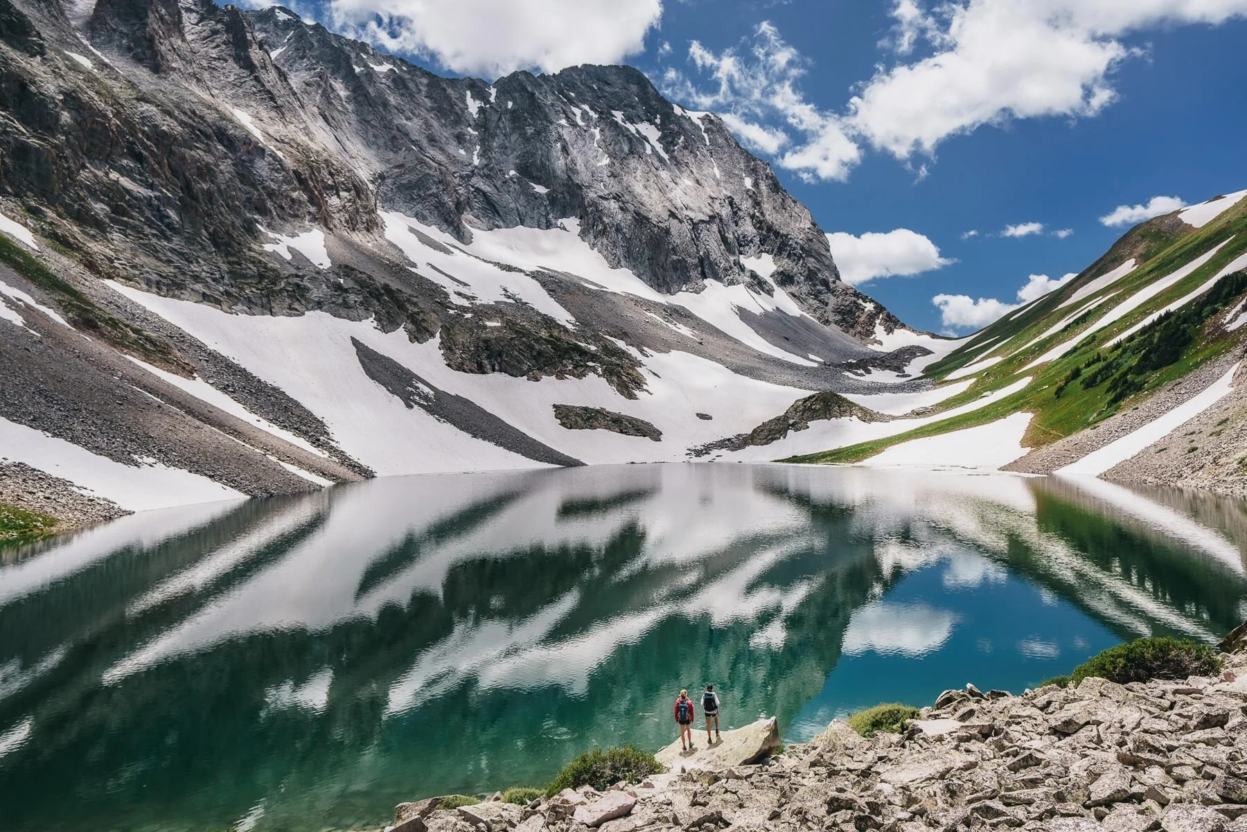 Two hikers with backpacks standing on rocks beside a calm mountain lake surrounded by snow-capped peaks and green slopes under a partly cloudy sky.