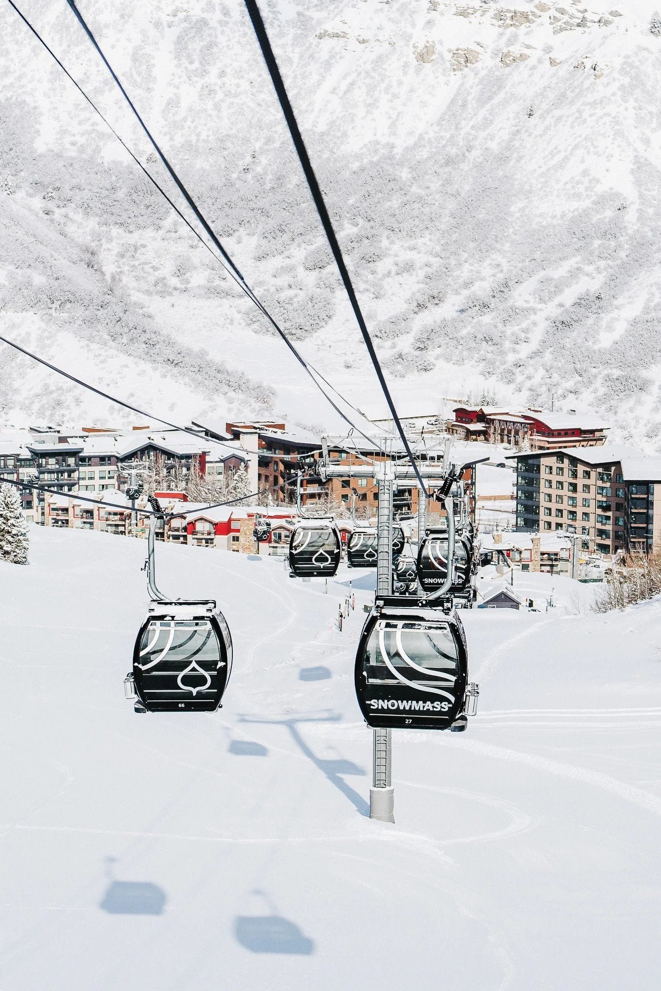Ski resort with snow-covered ground, ski lift chairs labeled 'SNOWMASS', residential buildings in the distance, and snow-covered mountains in the background.