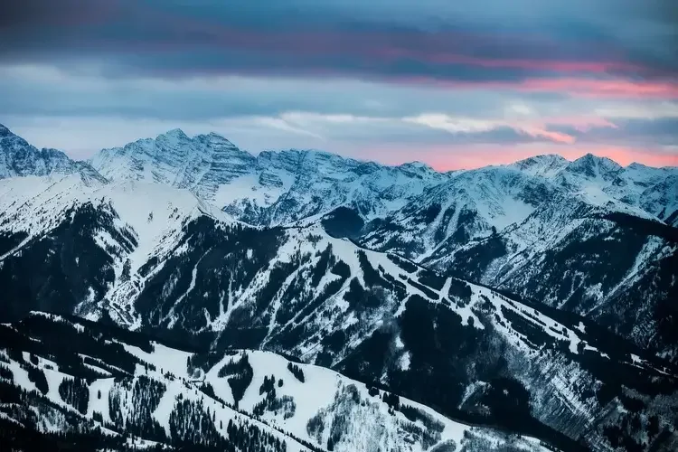 Snow-covered mountain range at sunset with colorful sky and dark clouds.

