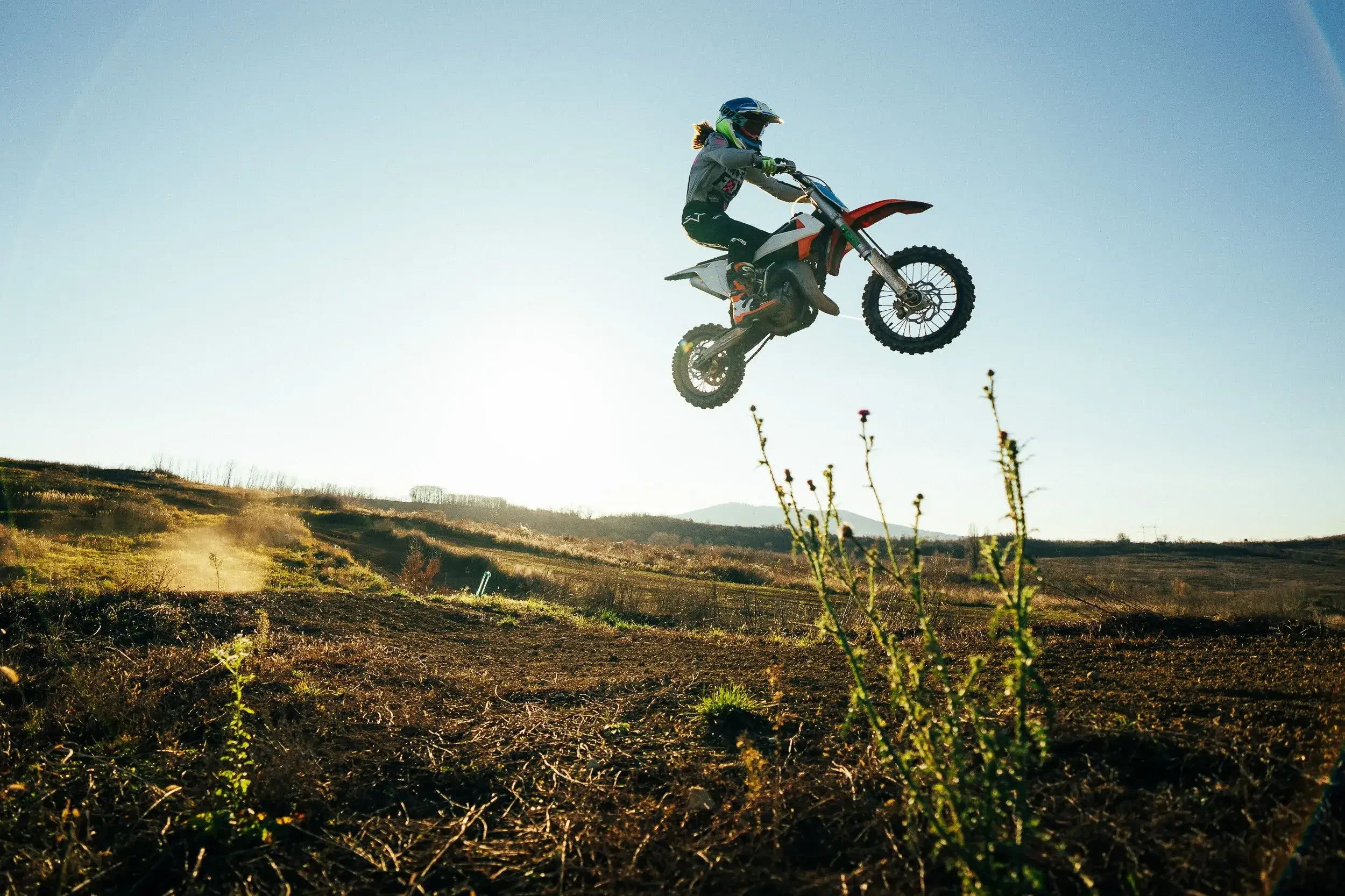 A motocross rider performing a jump in mid-air on a dirt track with the sun shining behind them and distant hills in the background.
