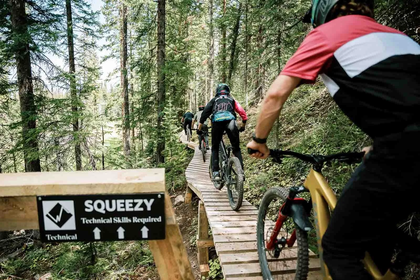 A group of mountain bikers riding on a wooden trail through a forested area.