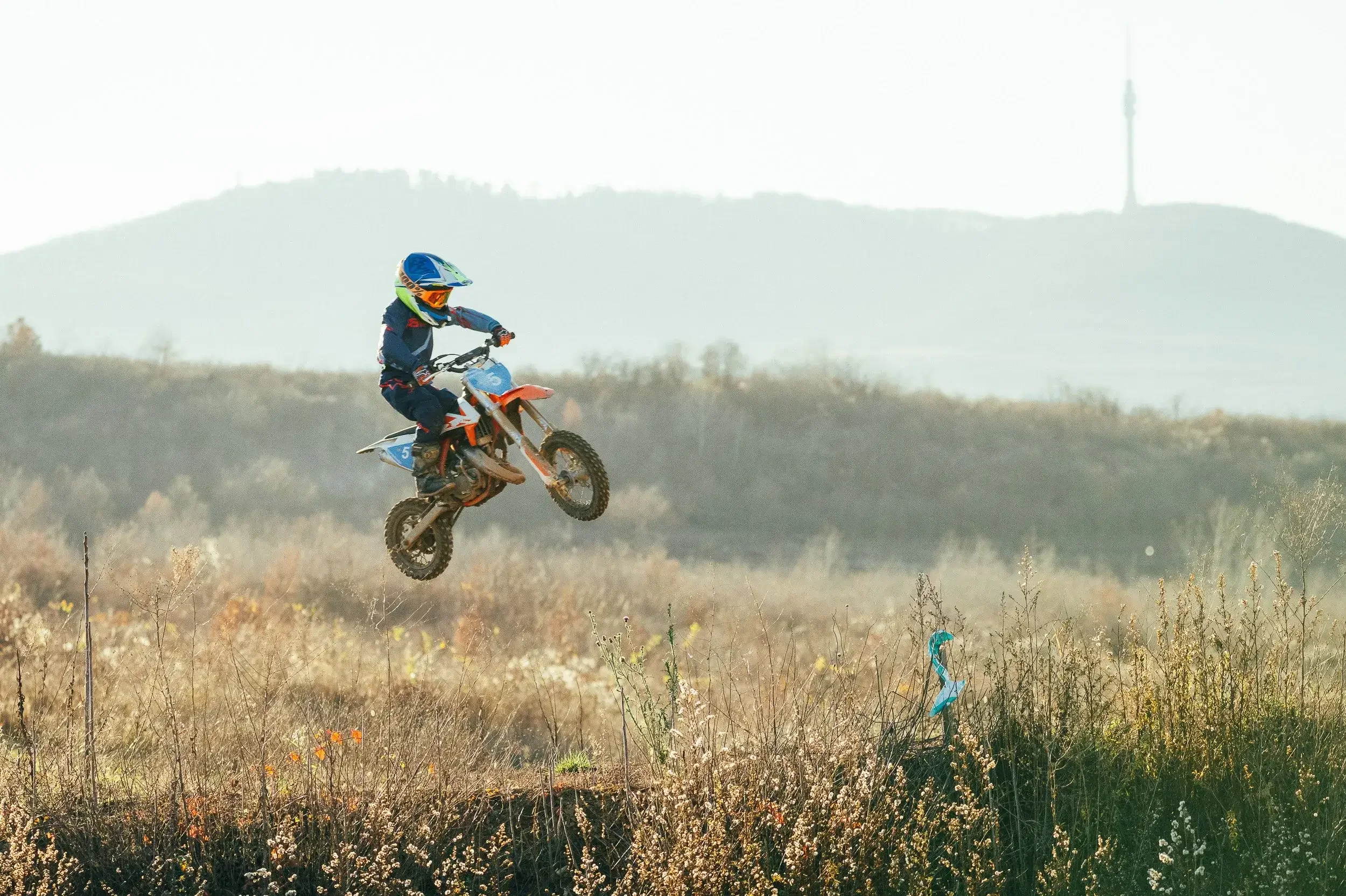 A young motocross rider wearing a blue helmet and racing gear jumps a dirt mound on an orange and white dirt bike in a grassy field with hills and a tower in the background.
