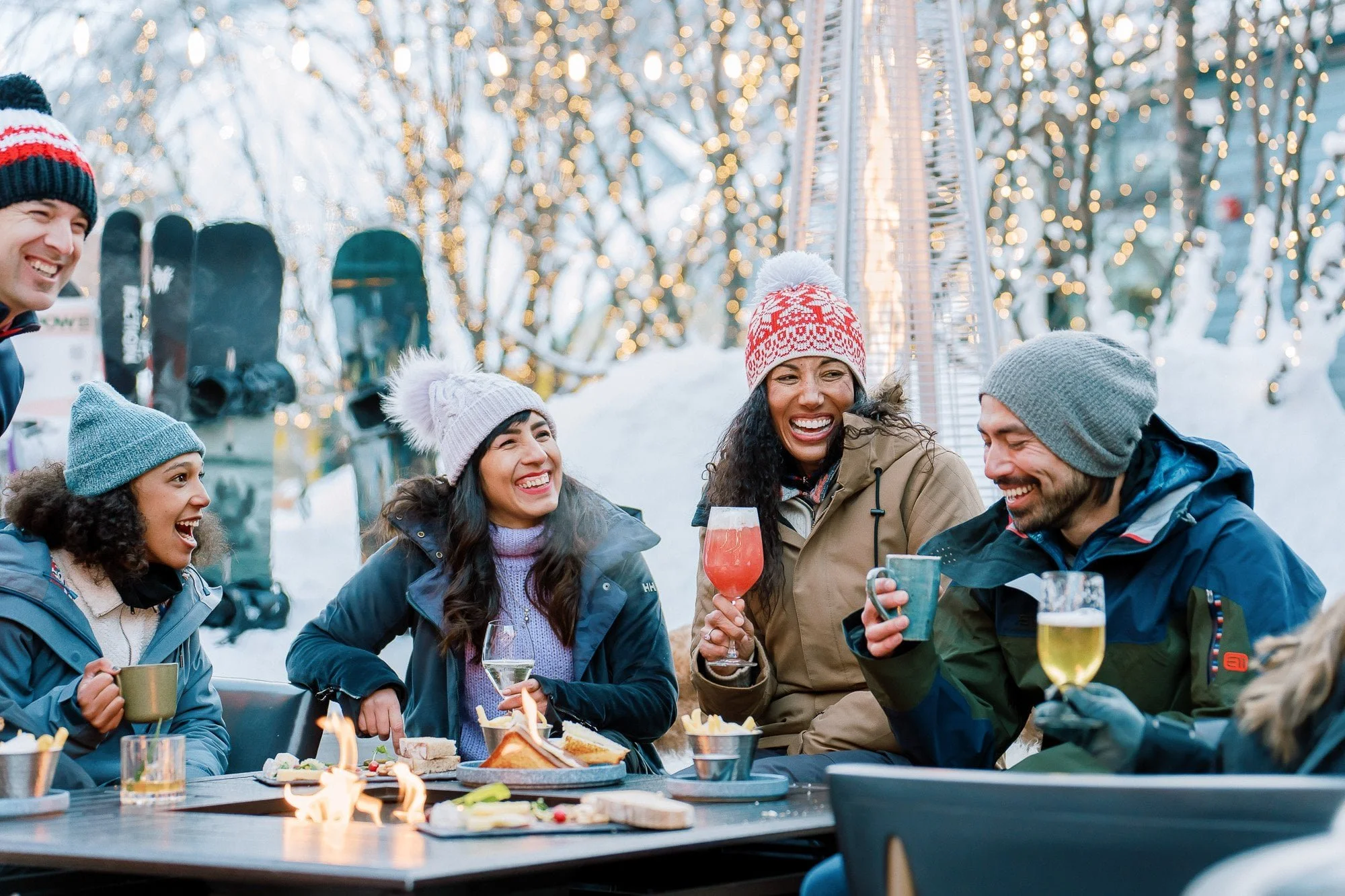 Group of friends outdoors in winter, wearing warm clothing and hats, enjoying drinks and food, with festive lights and snow in the background.