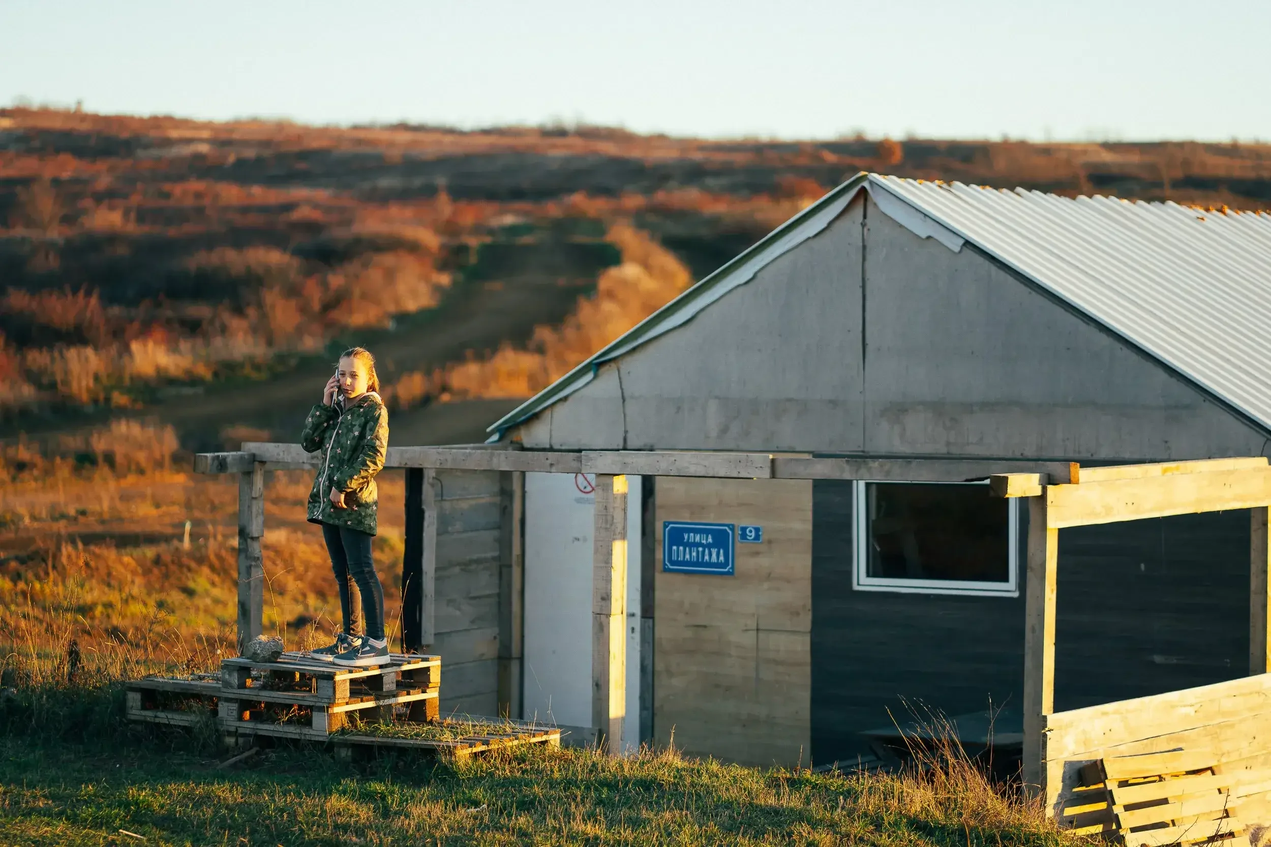 A young girl standing on a wooden pallet platform, talking on a cellphone, next to a house in a rural area with rolling hills in the background during sunset.
