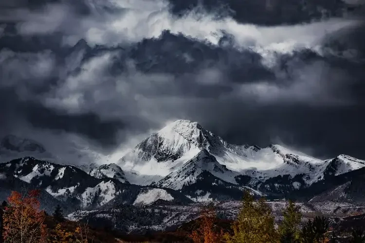 Snow-covered mountain peak under dark, cloudy sky with autumn trees in the foreground.
