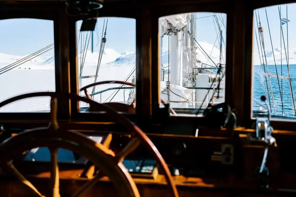 View from the helm of a boat showing snowy mountains and open water through windows.
