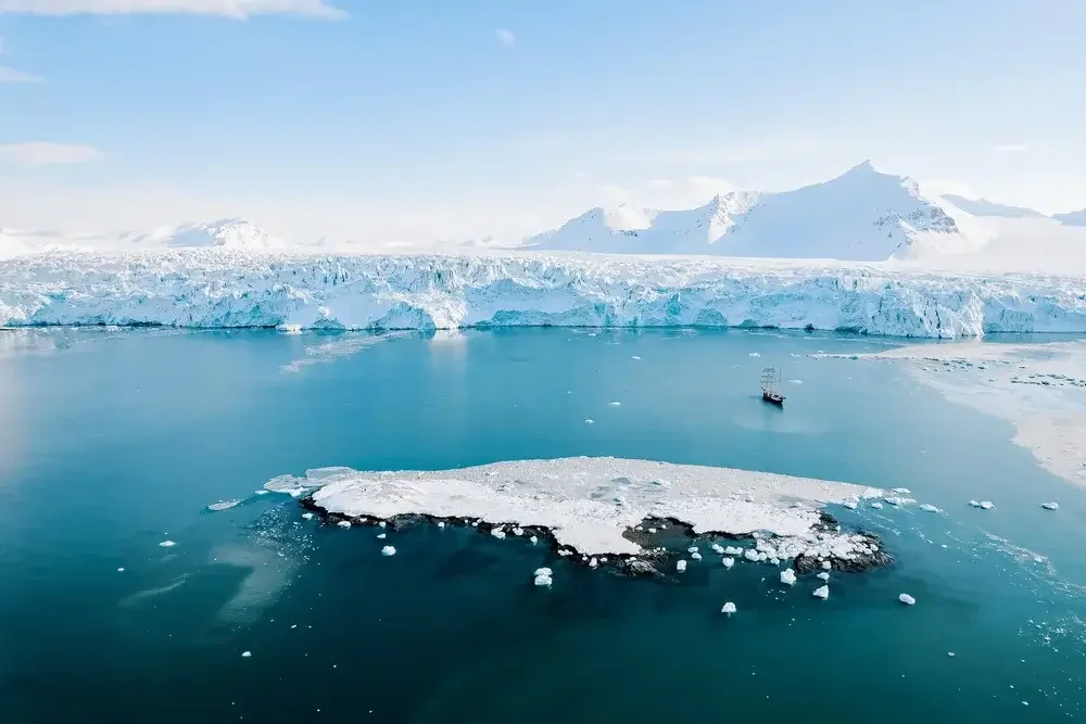 Frozen Arctic landscape with a glacier, mountain peaks in the background, a ship near ice floating in cold water.
