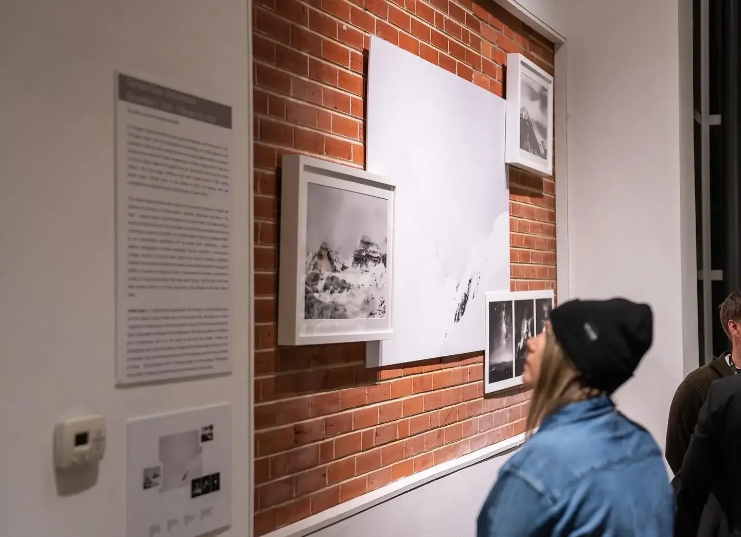 People viewing black and white photographs displayed on a brick wall in an art gallery.