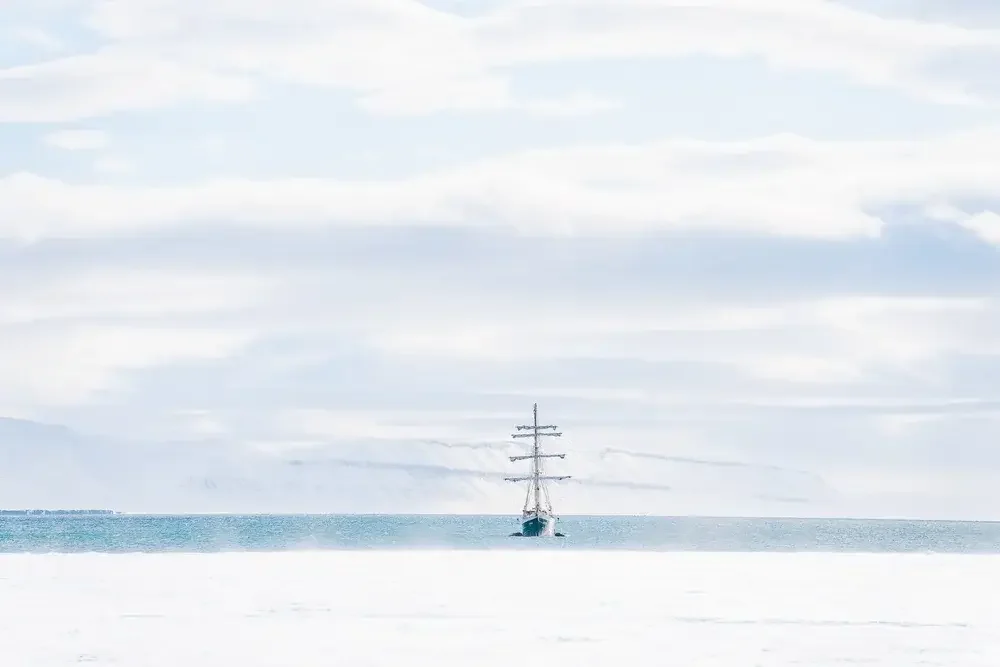A research vessel navigating through icy Arctic waters with crewmembers on deck, surrounded by fragmented ice and snow-covered mountains in the distance.
