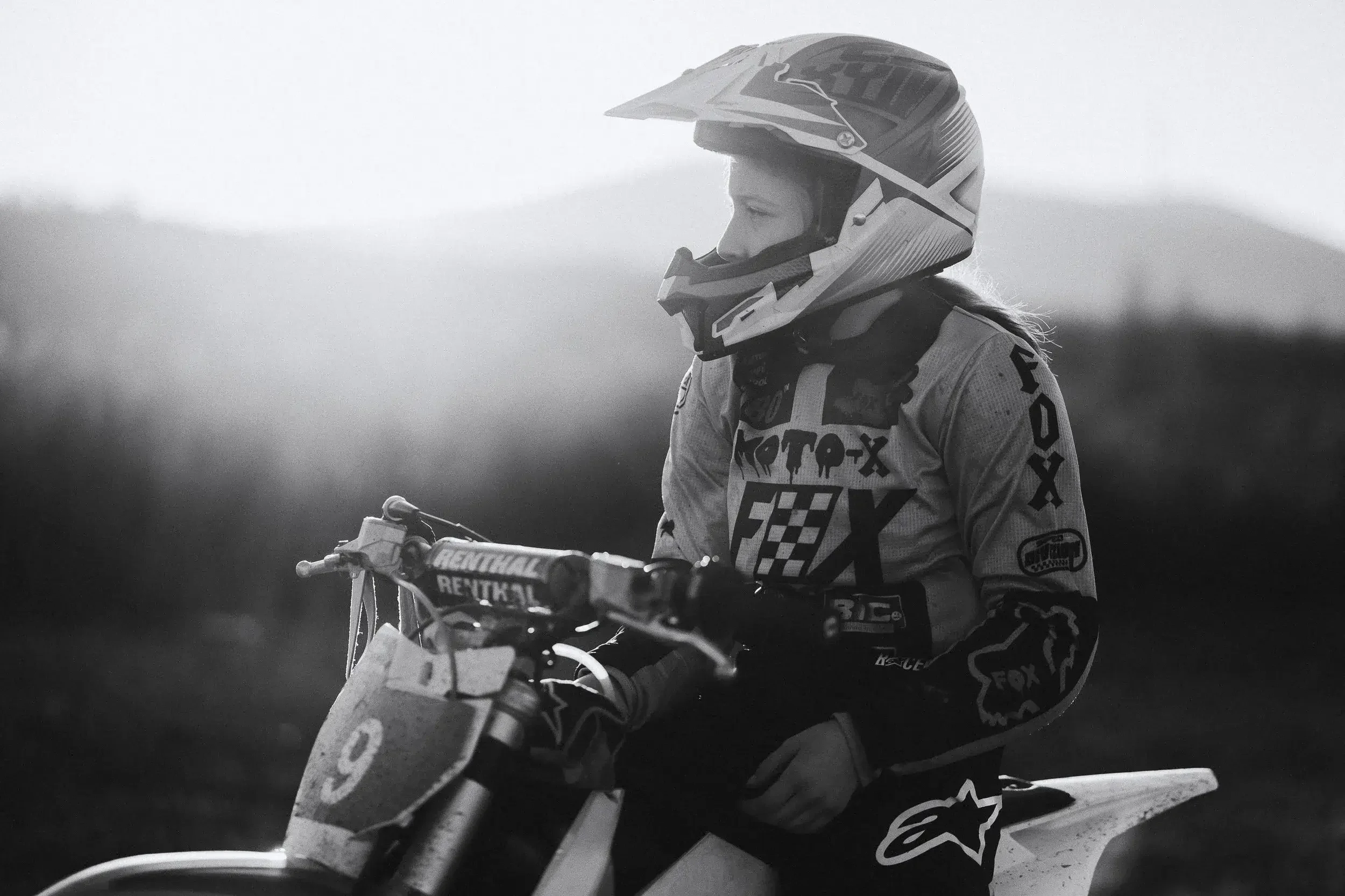 A female motocross rider wearing a helmet and racing gear sits on her dirt bike, looking to the right, with a blurred outdoor background.
