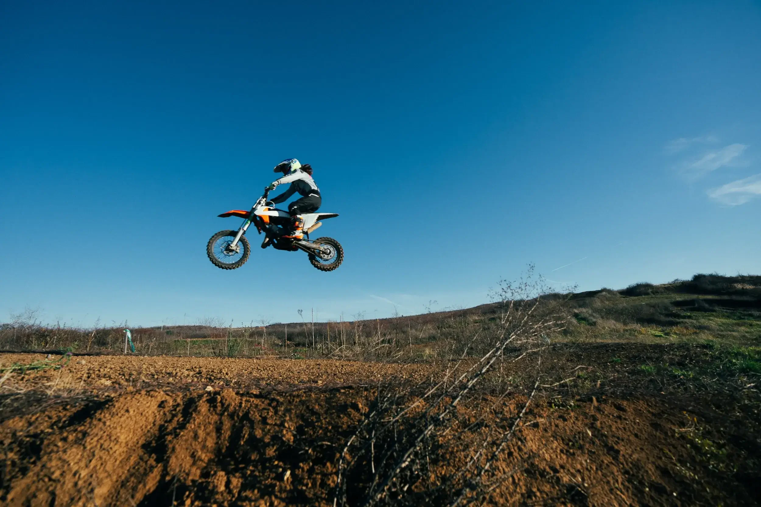 Motocross rider airborne on dirt track with blue sky background.
