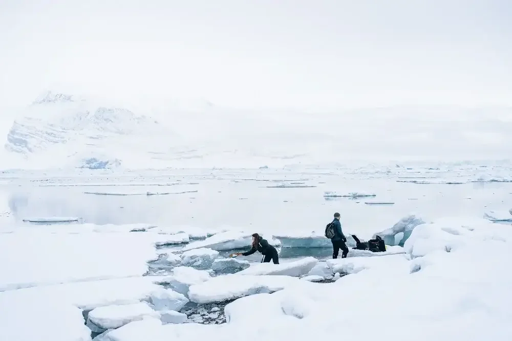 Two people in winter clothing exploring icy, snowy terrain near a body of water in a polar region, with mountains and icebergs in the background.
