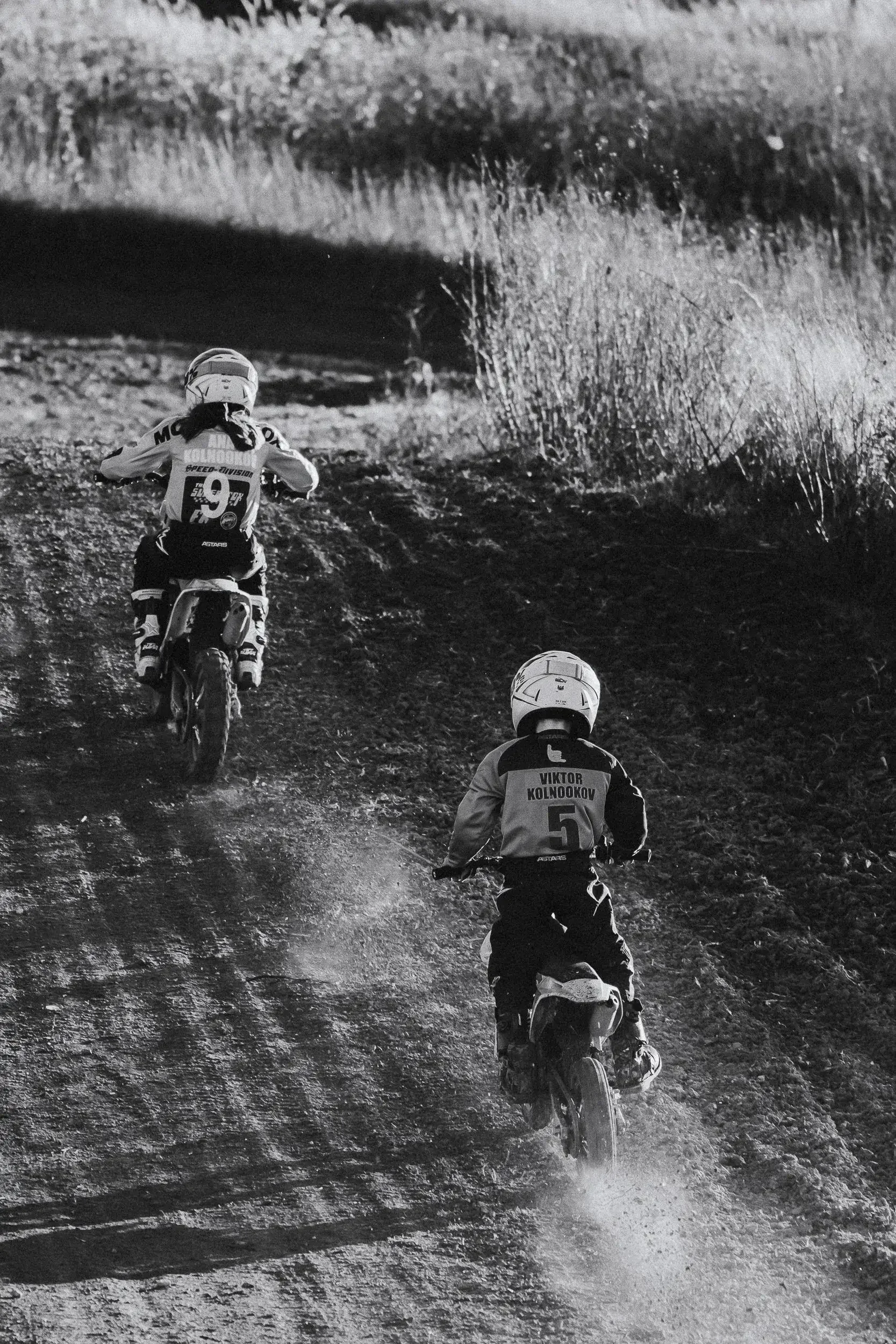 Two children riding dirt bikes downhill on a dirt trail in a natural outdoor setting.
