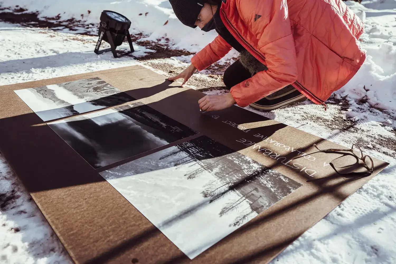 Person in a red jacket and black hat kneeling on snow, arranging black-and-white landscape photographs on a large brown sheet outdoors.