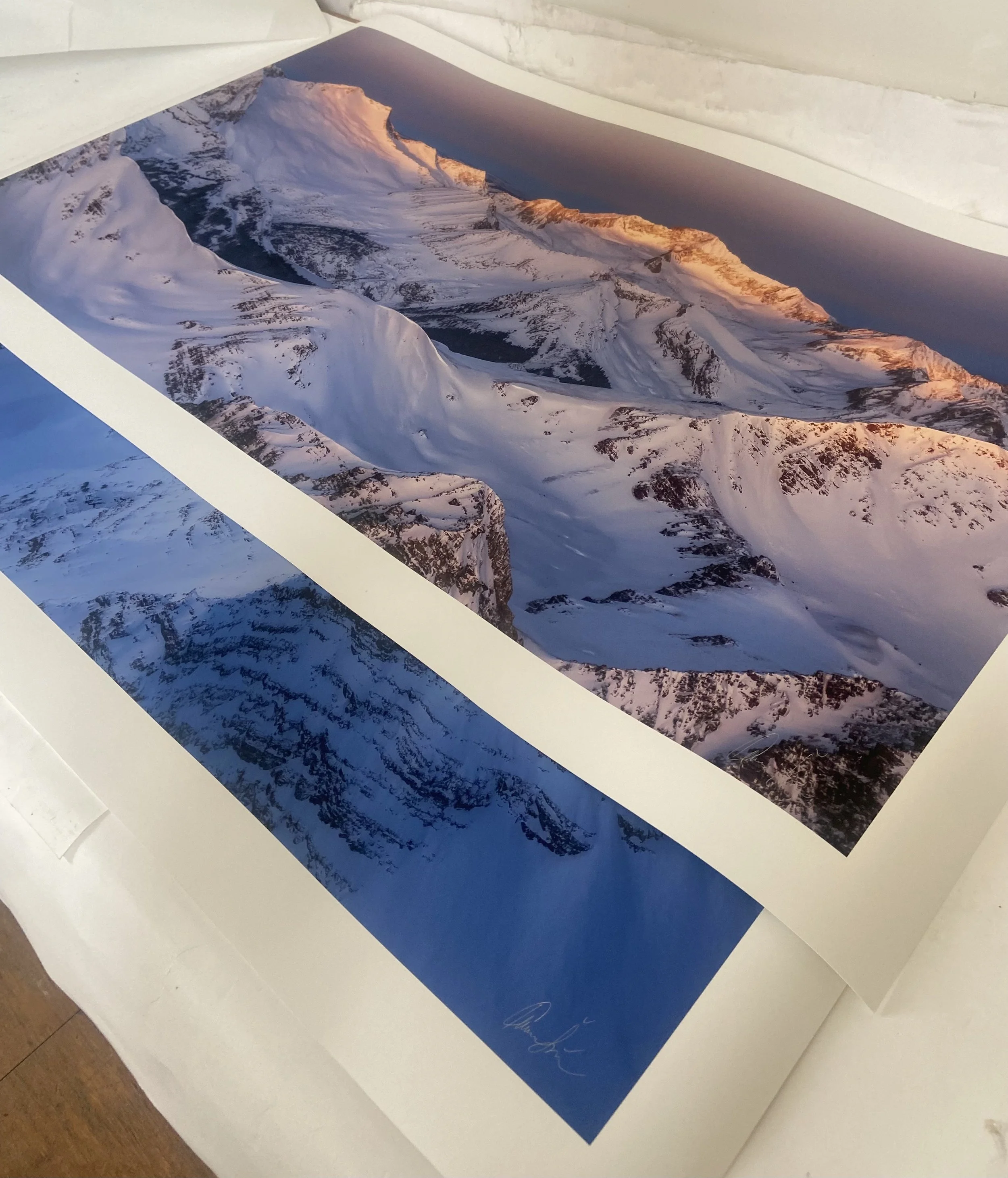 Print of a mountain landscape with snow-covered peaks and orange sunset lighting, set on a flat surface.