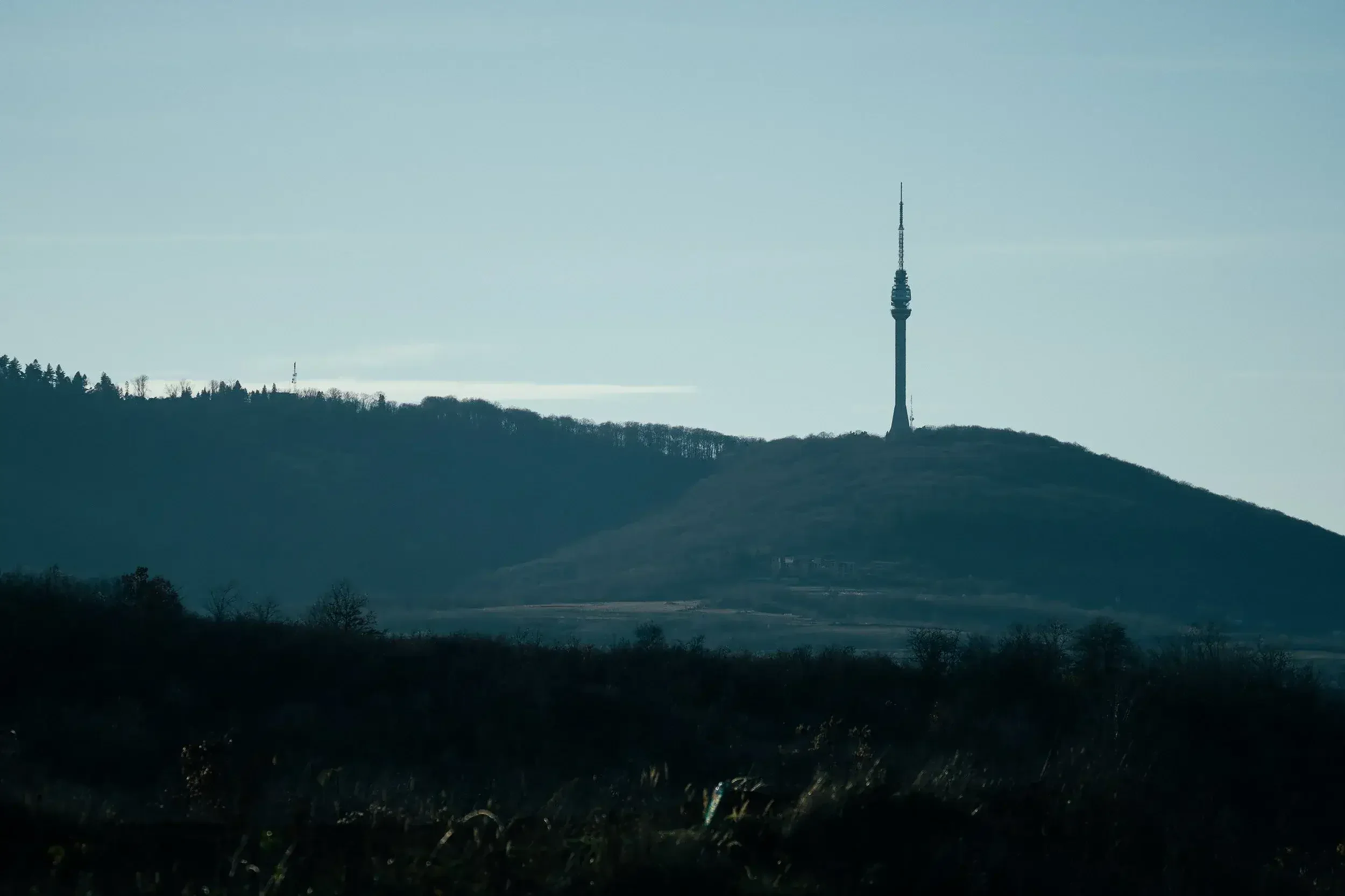A landscape of a hill with a tall tower on top, with distant trees and hills under a cloudy sky.
