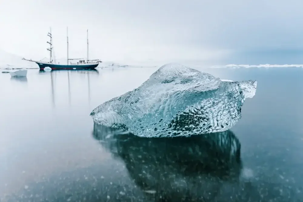 An icy landscape with a large irregularly shaped ice chunk in the foreground, calm water reflecting the ice, and a ship anchored in the background amid a snowy, overcast environment.
