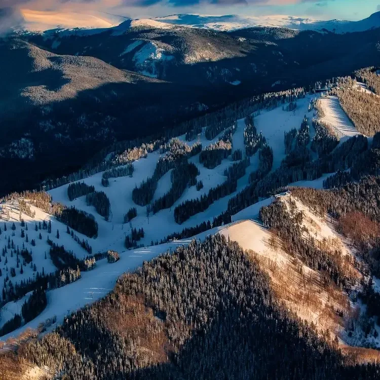 Snow-covered mountains and ski slopes in a forested mountain landscape during sunset or sunrise.

