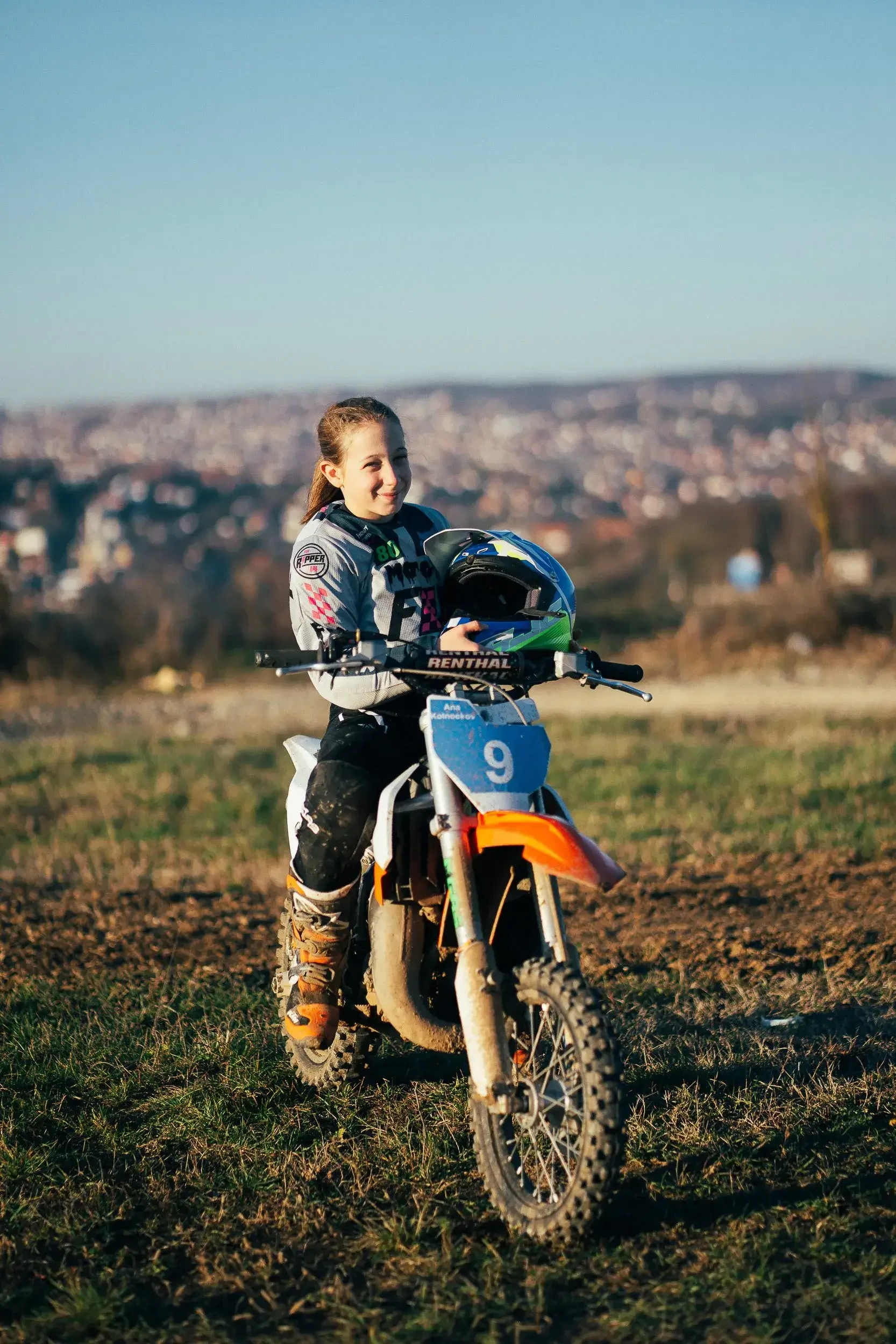 Young girl in racing gear sitting on a dirt motocross bike on a grassy field, smiling, with a cityscape in the background.
