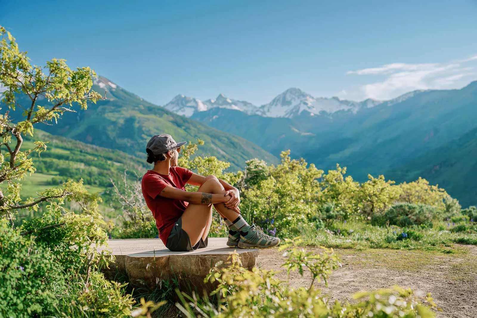 A person sitting on a wooden platform in a lush green landscape with mountains and snow-capped peaks in the background, wearing a hat, red shirt, shorts, and hiking boots.