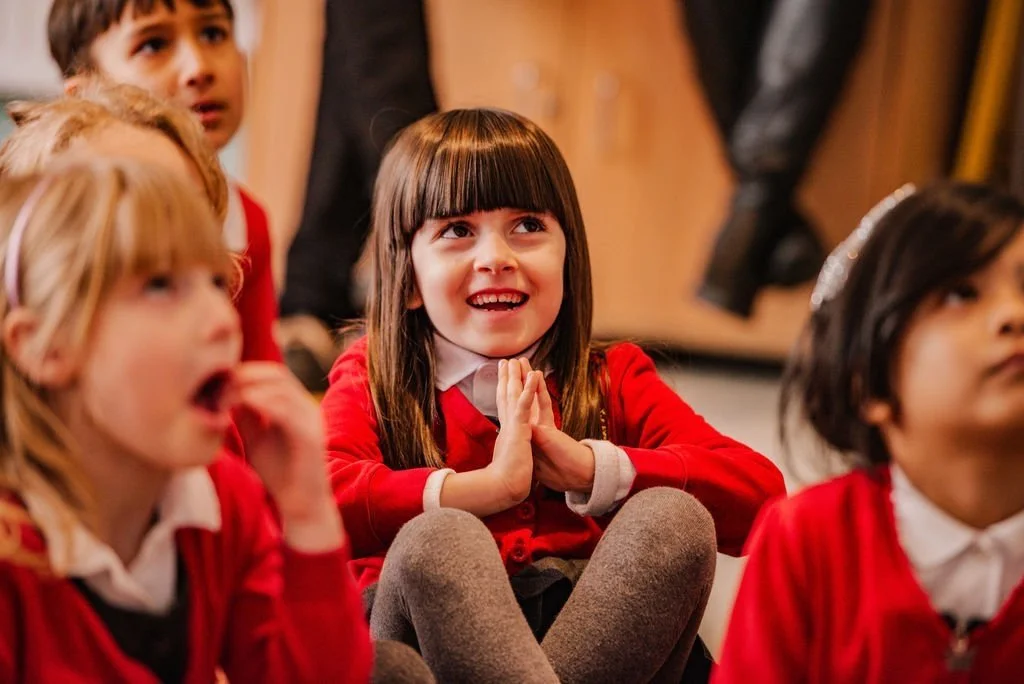 Children sitting and listening attentively in a classroom, wearing red school uniforms.