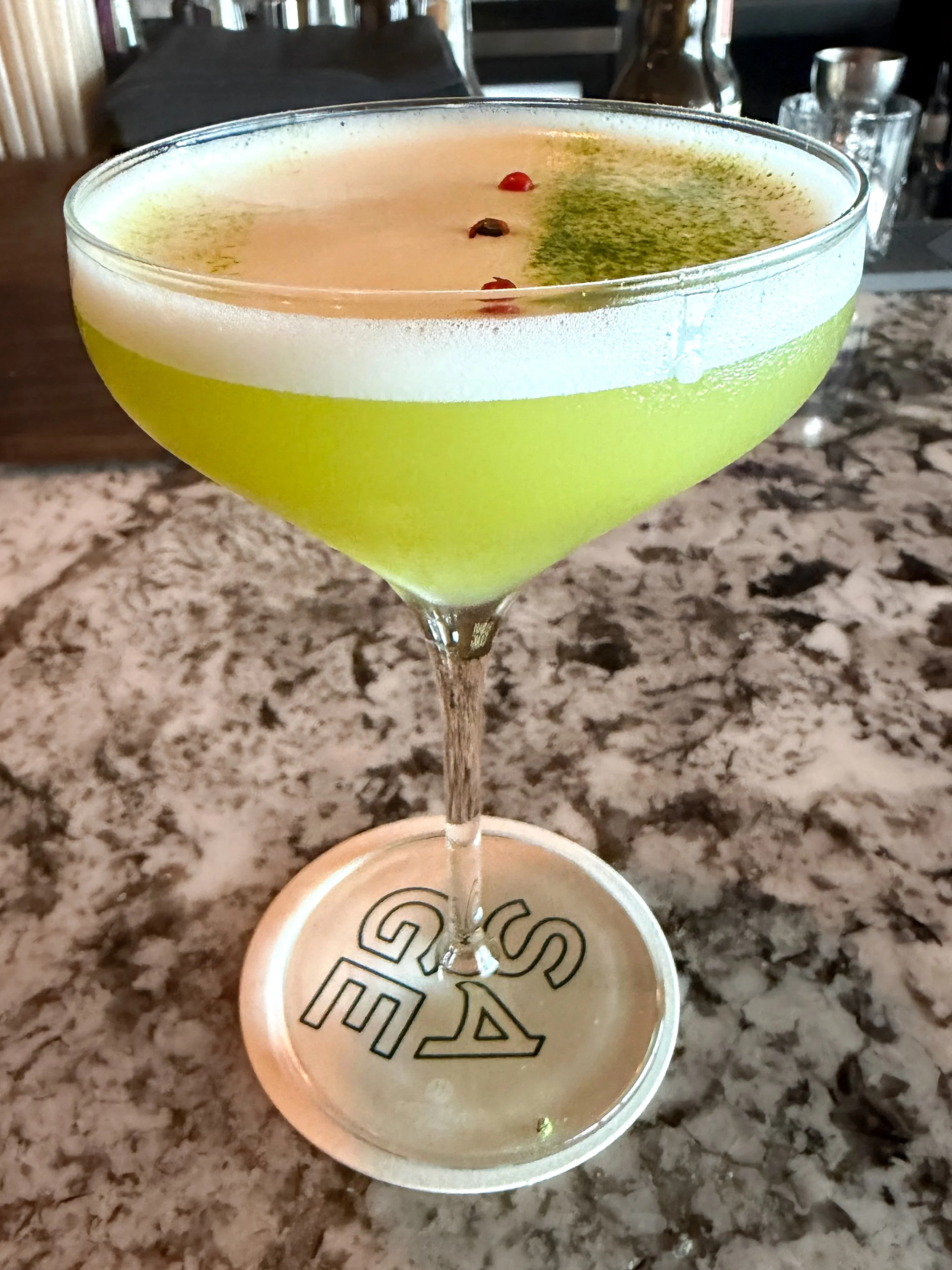 A green cocktail in a coupe glass with a frothy top, garnished with three small red berries and some green powder or herb on the foam. The cocktail is placed on a granite countertop with various glasses and bottles in the background.