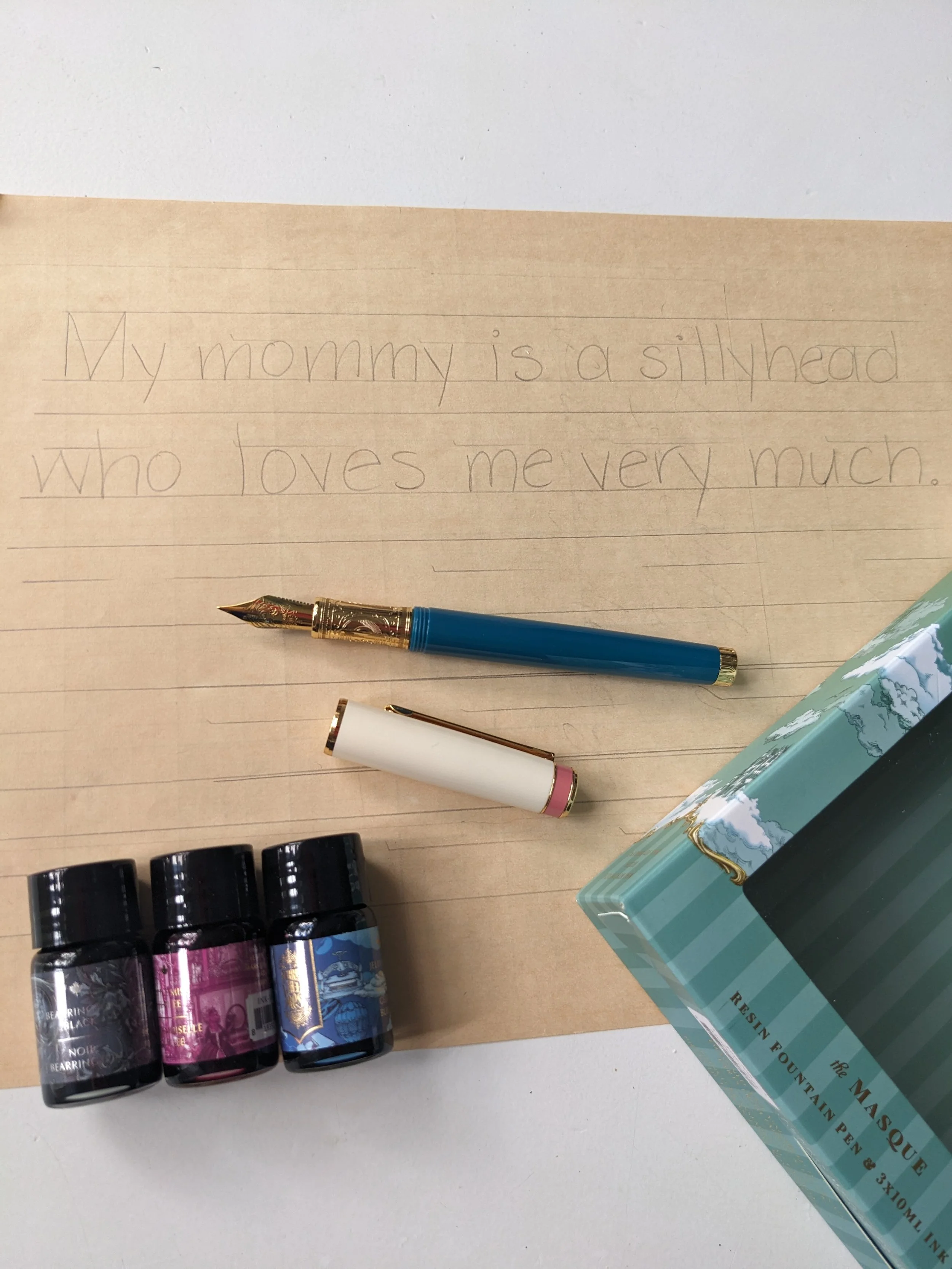 a sentence reading "My mommy is a sillyhead who loves me very much" on lined paper is next to a fountain pen and three small ink bottles
