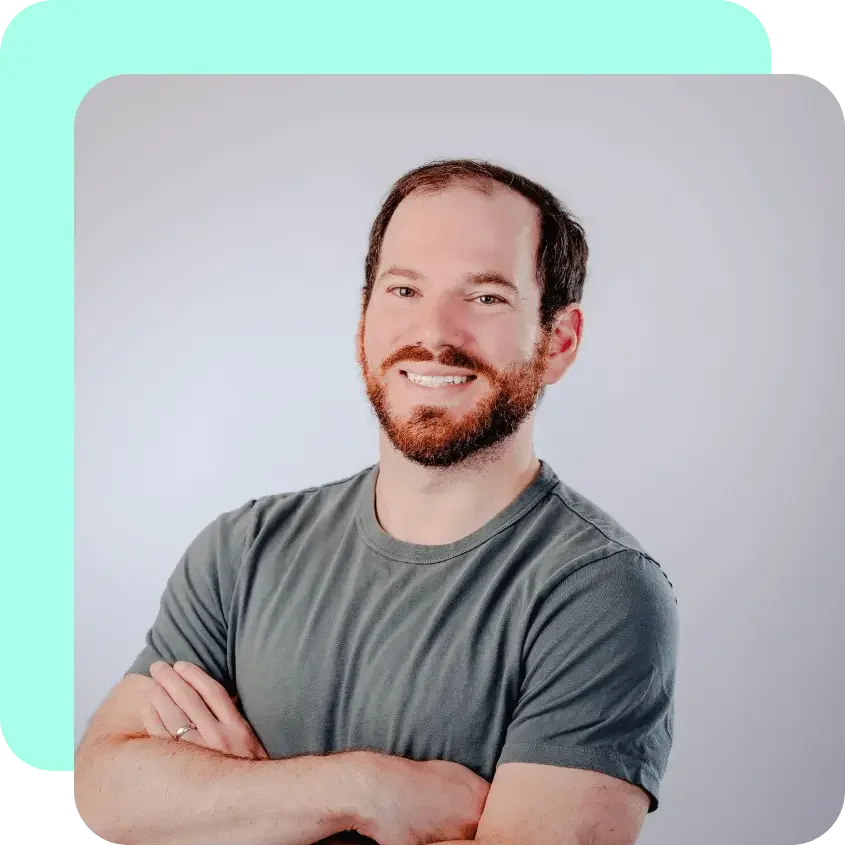 A smiling man with a beard and short hair, wearing a gray T-shirt, posing with arms crossed in front of a plain light gray background.