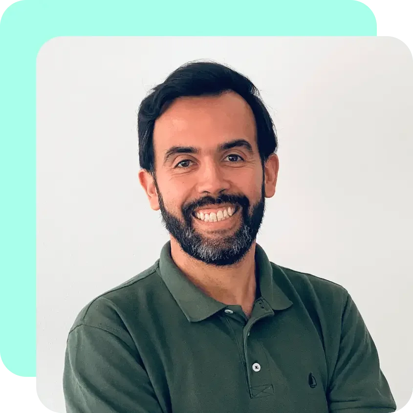 A man with dark hair and a beard smiling in front of a plain white wall, wearing a green polo shirt.