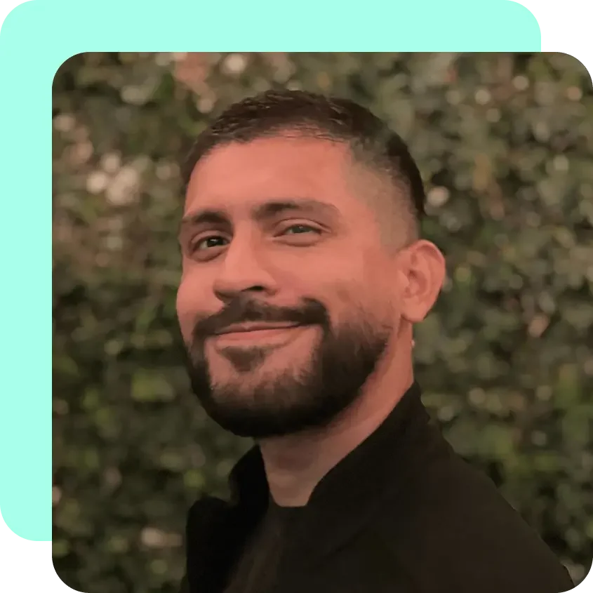A smiling man with a beard and short hair, wearing a black shirt, outdoors with blurred greenery in the background.