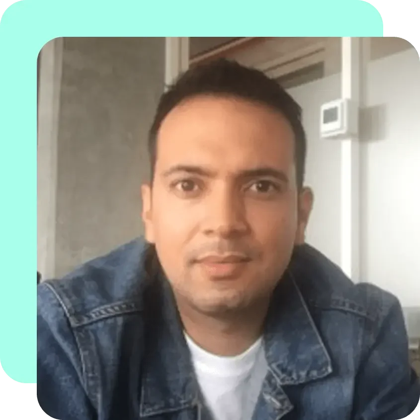 A man with short dark hair wearing a denim jacket and white t-shirt, looking at the camera in a modern indoor setting.