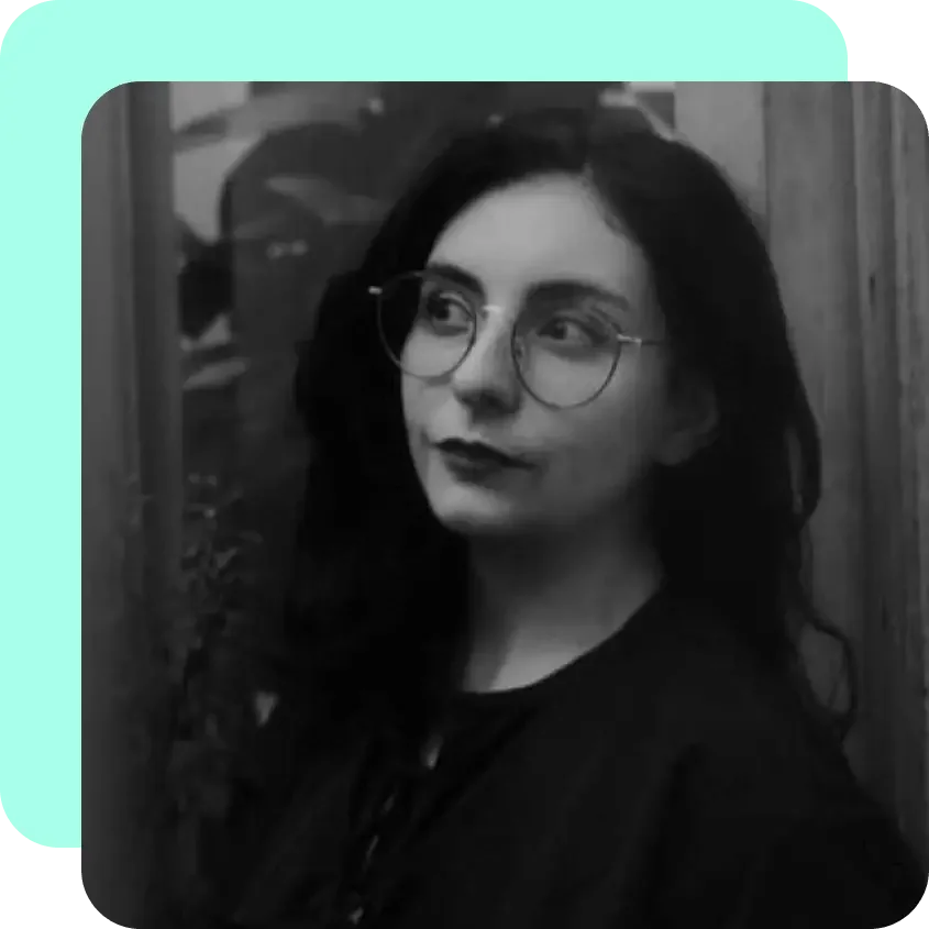 Black and white photo of a woman with dark hair wearing glasses and dark lipstick, standing outdoors near a wooden fence.