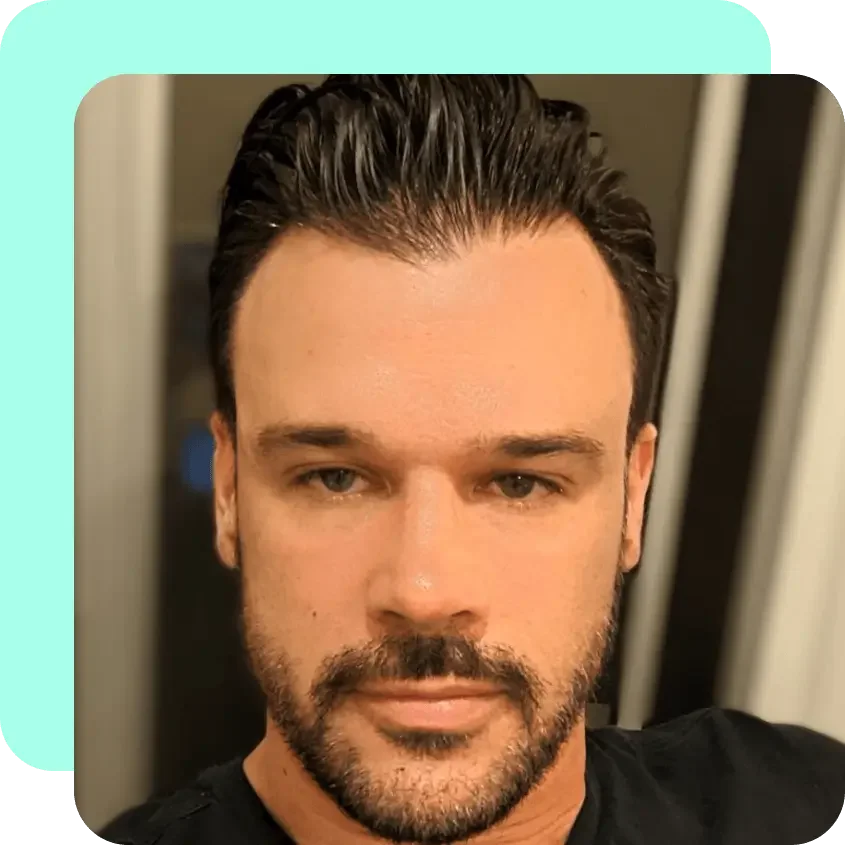 Close-up of a man with dark, slicked-back hair and a beard, looking at the camera indoors.